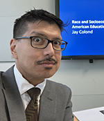 male with brown hair wearing tan suit and tie and glasses